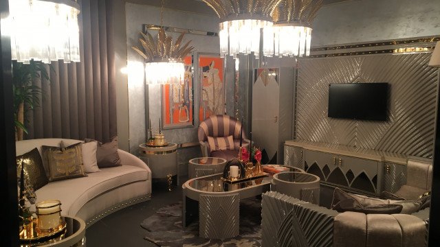This picture shows a luxurious living room with a mid-century modern style. It features a beige leather sofa, two armchairs, a low round glass coffee table, and a white shag rug. The walls are painted in a light grey color and decorated with art pieces and plants.