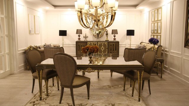 This picture shows a luxurious interior design that features a modern and elegant design. The room features a large cream sectional sofa with matching arm chairs, an ornate black and gold coffee table, glass side tables, and white tufted lounge chairs with beige and gold accent pillows. There is a sophisticated black and white rug and chandelier for lighting. Above, there is an intricately designed ceiling with painted white walls and gold trim. The overall look and feel of the room is grand and opulent.