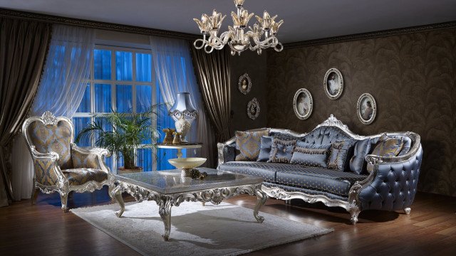 This picture shows a luxurious living room with a unique and modern design. The walls are white and feature two large abstract art pieces in blue, yellow and grey. The furniture includes a plush velvet couch, two armchairs upholstered in blue fabric and an ottoman with a glass top. A white chandelier hangs from the ceiling and a large area rug ties the space together.
