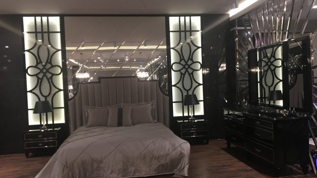 This picture shows a luxurious bedroom interior in a modern style. The walls and ceiling are white, with a dark wooden floor. There is an ornate bed with a gold-accented headboard and cream colored bedding. On either side of the bed are two sets of drawers for additional storage. On the wall behind the bed is a large mirror that matches the ornate frame on the bed. At the foot of the bed is a large seating area with a plush cream-colored sofa and a round glass-top coffee table. The overall look is elegant and sophisticated.