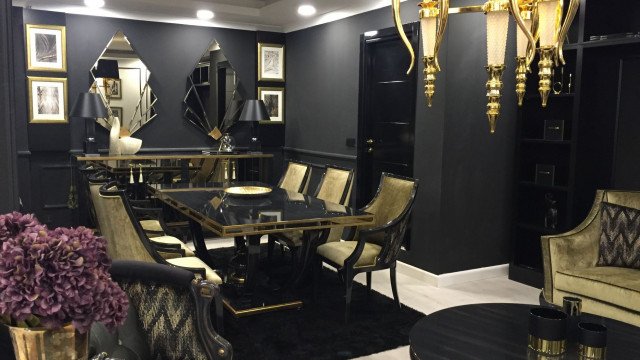 This picture shows an elegant, modern living room featuring a white and gold color palette. The room features comfortable seating with a white leather sofa and two armchairs upholstered in a black and white striped fabric. The walls are painted a soft white, and there is a decorative wall panel behind the seating area, providing texture and depth. A golden-colored coffee table and side tables complete the look, along with a vase of white and pink flowers. The overall look is bright and inviting.