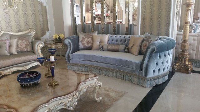 This picture shows an ornately decorated living room. The main feature of the room is a large curved white sofa, which is flanked by two end tables and two armchairs in shades of gold and brown. The sofa is set against a large staircase leading up to a balcony, and is decorated with pillows and throws in white, cream and gold colors. The walls are also adorned with a striped pattern wallpaper and a golden mirror. There is also a luxurious fur rug lying on the floor, which gives the room a very regal feel.