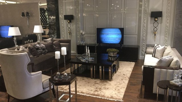 This picture shows a modern luxury living room decorated with elegant, classic furniture and accessories. The walls are a light gray color with white crown molding, and there is a large crystal chandelier hanging in the center of the room. The furniture is upholstered in cream colors with tufted details, and a plush area rug provides a comfortable place for seating. There are a few pieces of art hung on the wall, and the room is accented with gold accent pieces.