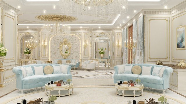 This image is a room design created by the interior design firm, Antonovich Design. The room features two cream colored armchairs upholstered in cream and gold fabric, with a matching sofa, all arranged around a mirrored accent table. The walls are painted an off-white shade, and the dark wooden floors contrast nicely against the neutral walls. A gold, sunburst mirror hangs above the fireplace, and deep red velvet curtains frame the windows. A white marble mantelpiece adds sophistication, and complements the intricate pattern of the carpet.