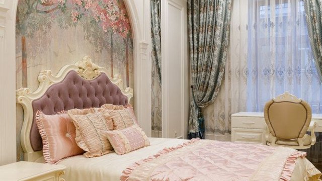 This is a picture of a luxurious bedroom interior featuring white walls and ceiling, a beige carpet, and traditional black furniture. A velvet armchair with a metallic gold frame stands in the corner, and a large leather bed with plush grey cushions and a patterned velvet blanket dominates the center of the room. Chandeliers and wall sconces light up the space, while tall curtains frame the windows.