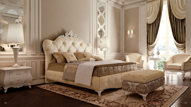 This picture shows a luxurious living room decorated with exquisite furniture and decor. The walls are painted with a light beige color and the floor is tiled with elegant marble tiles. A large, ornate crystal chandelier hangs from the ceiling, adding a touch of opulence to the room. The furniture consists of two plush sofas, an upholstered armchair, and a glass-top coffee table. Several throw pillows, plants, and accessories complete the look.