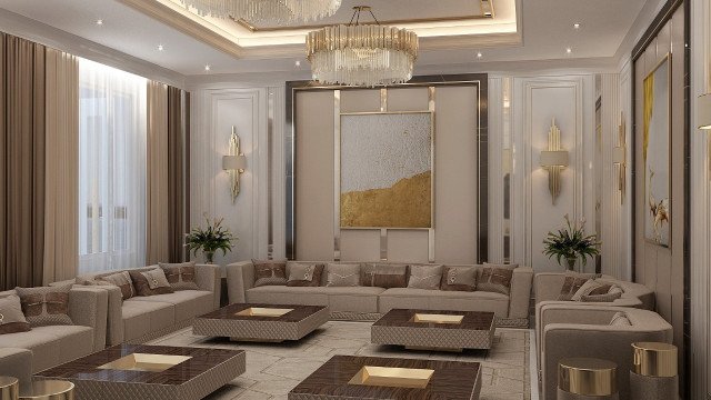 The picture shows a luxurious and modern living room, with an expansive white sectional sofa and armchair set up around an opulent gold and marble coffee table. The walls are painted in a classic cream color, and the room is accessorized with large pillows, a grand chandelier, and floor-to-ceiling sheer curtains framing the tall windows. The parquet wooden flooring completes the look to create a sophisticated space for relaxation and entertaining.