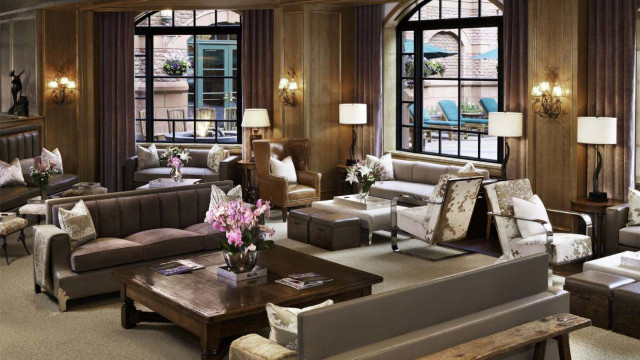 The picture shows a luxury open-plan living room with a white and gold theme. The walls are painted a light color and the furniture is made of white leather and white marble. There is a large chandelier hanging overhead, and gold details throughout the room, including on the frames of the large windows and the end tables. An elegant curved staircase is situated in the middle of the room and leads to an upper floor.