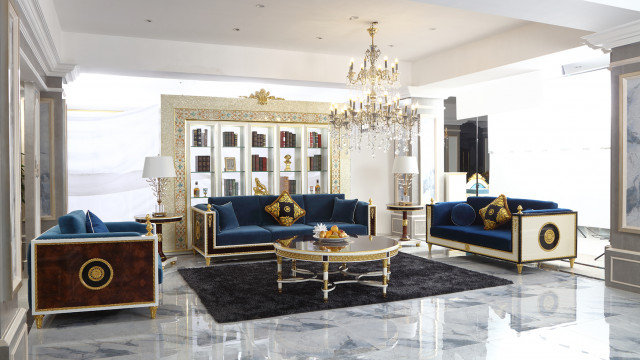 This picture shows an elegant living room interior design, with high-end furniture and decor. The walls are painted a warm shade of beige, while the floor is covered in a white marble pattern. The room has two sofas upholstered in light gray fabric, along with several armchairs with matching gray cushions. There are two end tables on either side of the room, one featuring glass vases and a decorative lamp and the other with a silver tray and a bowl of fruit. In the center of the room is a woven area rug with a geometric pattern in shades