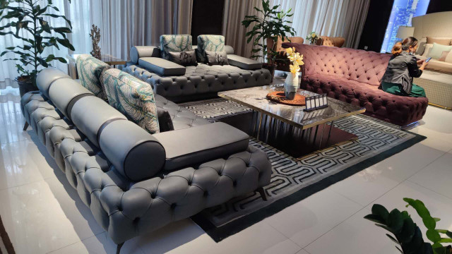 This is a picture of a modern living room interior design. The room has a light blue and beige color scheme, with grey accents throughout. It features a seating area with a dark blue velvet sofa and two armchairs, as well as a coffee table and a large rug in the center. There is a built-in television unit on one wall with a marble top and two pendant lamps above it. On the opposite wall is a wall-mounted fireplace with a mantel, surrounded by bookshelves and storage cabinets. The room also includes several floor-to-ceiling windows