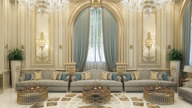 This picture shows a luxury living room with a white and gold color palette. The room has a large, comfortable sofa in the center with two armchairs facing it. On the right side, there is a television mounted on a wall and a fireplace with a white mantle. The space is bordered by a large Persian rug and features a round coffee table with a white marble top and accents of gold throughout. The ceiling features recessed lighting, adding to the overall luxurious feel of the space.