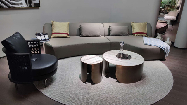 This picture shows a modern and luxurious living room interior, designed by Antonovich Design. It features a contemporary white sofa, adorned with several gold-accented throw pillows, as well as a white, tufted ottoman. The walls are painted dark grey, and there is a large grey velvet area rug in front of the sofa. A round metal and glass coffee table with two matching armchairs are also featured in the room, while two tall, stylish lamps create an ambient lighting. Finally, some potted plants bring a relaxing and natural touch to the space.
