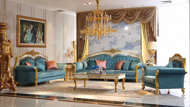 This is a photo of a modern and luxurious living room. The walls are painted a light grey, with abstract art decorating the walls. There is a white sofa and ottoman against the wall, with patterned ivory and gold cushions. Several accent chairs surround a large glass coffee table filled with decorative books and objects. The floor is covered with a large white shag rug and there are pendant lights hanging from the ceiling, adding a touch of luxury.