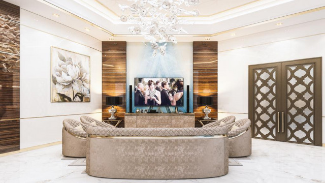 This picture shows a luxurious modern home interior. The room features two large leather chairs and a sofa arranged around an elegant stone coffee table. The room is illuminated by several lamps and a grand crystal chandelier, and the floor is covered in a rich cream-colored carpet. On the walls, various pieces of art hang in bright gold frames, adding to the ambiance of opulence.