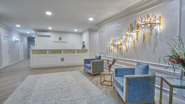 This picture shows a modern and luxurious living room design. It features a white marble floor, an intricately patterned rug in the center, two comfortable grey sofas, velvet armchairs, and a large crystal chandelier hanging from the ceiling. There is also a built-in white fireplace with a rustic wooden mantelpiece, while the walls are covered in a rich mahogany paneling. The room is illuminated by recessed lighting and several windows along the back wall.