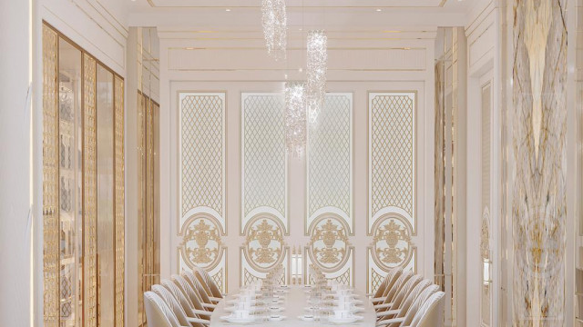 This picture shows a luxurious interior design in a grand hallway of a wealthy home. The walls are covered in a white and beige marble pattern, with a large stone mantelpiece at one end. In the center of the hallway is an ornate chandelier hung with several rows of crystal beads. The floor is composed of a black and white checkered marble pattern. Two small alcoves are seen on either side of the hallway; one with a decorative potted vase, and the other featuring a marble statue. An elegant set of double doors can be seen at the