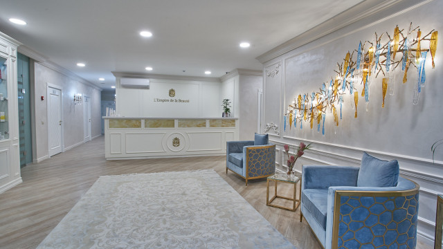 This picture shows a modern living room design. The room has a blue, white, and gold color palette. There are two beige sofas with white and gold cushions, a gold round coffee table, a beige armchair, and a white and gold TV console in the center. The walls are decorated with overlapping abstract paintings in shades of blue and a gold mirror. There are several potted plants around the room to add a touch of greenery and a white luxurious chandelier hanging from the ceiling.