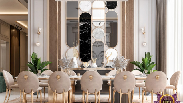 This picture shows a luxurious dining room with a large, two-tiered ceiling, ornate chandeliers, and recessed lighting. The walls are decorated with wood-paneled wall features and gold accents that are complemented by the white and grey marble flooring. The centerpiece of the space is a long, rectangular dining table with white upholstered chairs and intricate accents on the walls. An elegant, curved staircase can be seen in the background, leading to the second level of the room.