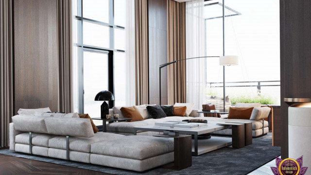 This picture shows a luxurious living room designed by Antonovich Design. It features a large sectional sofa upholstered in a velvety dark grey fabric, with a coordinating grey tufted ottoman and several comfy accent chairs. The walls are painted a light gray and are adorned with modern wall art and a bold black chandelier. The floor is covered with a plush, beige shag area rug, and the room is illuminated with recessed lighting and wall sconces.