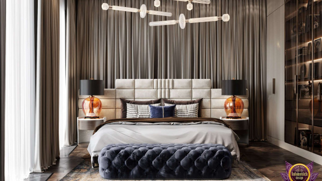 This picture shows a modern luxury living room with high vaulted ceilings, a large white sofa with plush gray and white pillows, a glossy black coffee table with a glass top, and a unique antique chandelier as the main focal point. The walls are a soft gray color with white trim, and the floor is covered in a light-colored carpeting. To the left of the sofa is a large window with a sheer white curtain, and to the right is a wall-mounted fireplace with a matching marble surround.