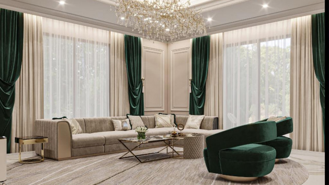 This picture shows an interior design featuring a luxurious and modern living room. The walls are painted in a light cream color with a dark brown accent wall. A large crystal chandelier is hung from the ceiling, and a grey sectional couch is placed in front of the accent wall. On the side of the couch is a glass coffee table with a decorative rug underneath. Above the accent wall is a large abstract painting and there are two white armchairs with metallic accents. On the other side of the room is a gorgeous marble fireplace with a wooden mantel.