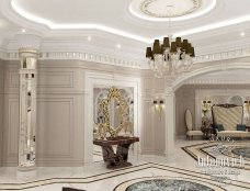 This picture shows an exquisite modern-style foyer with a golden tone. There is an elegant gold paneling throughout the walls with intricate marble mosaic on its floor. The ceiling of this foyer is also gorgeously crafted with elegant gold chandeliers and statement pieces. In the center of the foyer, a metallic round table has been placed with a vase of pink calla lilies. The furniture pieces feature sleek and luxurious accents that add a luxurious touch to the overall design.