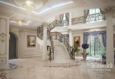This picture is of a large elegant foyer in an aparment building. The floor is marble and the ceiling has a beautiful chandelier and other ornate details. There is an inviting curved staircase with a large window at the bottom which allows for lots of natural light to come in. The walls are a neutral color and have sconce lighting fixtures and some artwork hung on them for decoration. The room is large and open, making it perfect for welcoming guests.