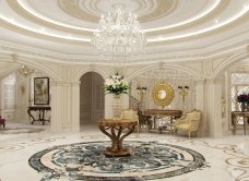 This picture shows a luxurious formal living room with white walls, a marble floor, and a large crystal chandelier. There are several plush upholstered couches and chairs in a cream and beige color palette. Decorative accents such as a round gold mirror, statue busts, and white flower arrangements add elegance to the space. A built-in bookshelf adds character and a warm, inviting ambience.