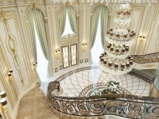 This picture shows a luxurious interior design with marble flooring and walls, and a grand staircase made of white marble. The walls are adorned with beautiful art pieces and the staircase features intricately carved balustrades. A large chandelier hangs from the ceiling and the room is illuminated by floor-to-ceiling windows that also provide a stunning view of the outdoors. The overall effect is one of grandeur and luxury.