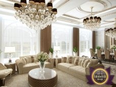 This picture shows a luxurious living room with a high vaulted ceiling, white walls and marble floors. The room is furnished with creamy furniture, a cozy velvet armchair and ottoman, a large bookshelf, and a marble fireplace. The walls are decorated with intricate gold frames, patterned curtains, and a large round mirror. There are several tall windows that allow natural light to shine in, adding to the comforting atmosphere of the room.