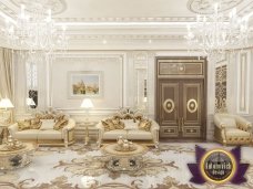 This picture shows a modern interior design with a white and gold color scheme. The walls and floor are covered in white marble and there is an intricately carved white marble fireplace in the center of the room. There is a large black sofa with gold accents and a white ottoman in the center of the room. To the side is a gold and white abstract painting hung on the wall, and a contemporary gold coffee table sits underneath. The lighting coming from the chandelier gives the room a warm and inviting ambiance.