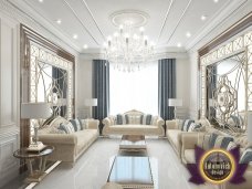 This picture is showing an interior design project from the luxury design firm Antonovich Design. It features a living room with a luxurious off-white sofa, an oval coffee table, and a large floor rug. The walls of the room have been decorated with wall panels and decorative frames, and the ceiling has an intricate pattern of lights arranged in a star-like shape. There is a dark wooden cabinet in the corner of the room, and the windows are dressed with white curtains.