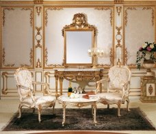 This picture shows a luxurious bedroom design decorated in white and gold tones. The room has a high ceiling with intricate gold-trimmed details. On the walls there are two large gold-framed art pieces. The bed is a four poster bed frame, with a plush white headboard, and a white and gold silk canopy draped above. The bedding is white and silver, while golden accessories add accents of elegance. The bedside tables and carpeting have both been chosen to match the overall decor of the room.