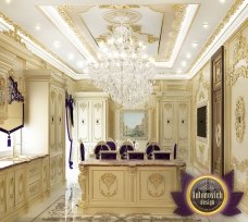 This picture shows a luxurious white dining room with a large, dark wooden table in the center, surrounded by eight white chairs. The walls are decorated with white crown molding and gold sconces. The room is lit with a beautiful crystal chandelier, adding a sense of grandeur to the space. There is a large window in the corner, allowing natural light to enter the room.