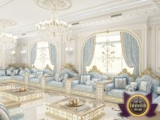 This is a modern luxury living room with white walls and furniture. There is a large grey sectional sofa in the center of the room, with a glass coffee table in front of it. The walls feature art pieces in gold frames, while two matching side tables sit against one wall. There is an ornate floor lamp in the corner, and a crystal chandelier hanging from the ceiling.