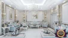 This picture shows a luxurious white and gold living room with a grand fireplace at the center of the room. There are intricate carvings on the fireplace mantel, paired with two large chandeliers that hang from the ceiling. On either side of the fireplace there are two dark wood sofas that have golden accents, as well as a matching set of armchairs. The walls and floors in the room are also white, with a brown and beige patterned area rug lying in front of the sofa. Gold-framed art and ornate decorations complete the look.