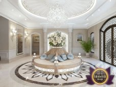 This picture shows an elegantly designed modern hallway with white marble floors, gold and white walls and ceilings, and a light gray couch with gray and white cushions. A stunning and luxurious chandelier hangs over the couch, adding a touch of glamour to the space. The hallway is decorated with various artwork and several stylish wall sconces. On either side are two sets of double doors that lead into other parts of the home.