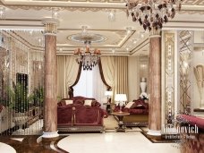This picture shows a large, luxurious hotel lobby. It has a high ceiling with intricate decor including several chandeliers, two grand staircases, and ornate columns and furniture. The walls are paneled in dark wood, and the floor is marble with a beautiful geometric pattern. In the center of the room is an eye-catching sofa and armchairs set around a low coffee table. There is a large decorative fireplace to the left and two large potted plants near the staircase.