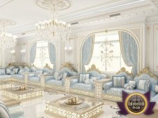 This picture shows a luxurious living room interior design. The room is decorated in an elegant and sophisticated style, with plush cream-colored furnishings, lots of patterned pillows, and a large ornate area rug providing a dramatic focal point. Creamy white walls are set off by tall gold-trimmed windows and a large chandelier hanging down from the ornately carved ceiling. The built-in cabinet provides additional storage and decorative accents complete the look.