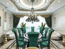 This picture shows an interior design concept for an opulent dining room. The walls are painted in a light gray color and feature ornate gold patterned wallpaper along the top half of the wall. In the center of the room is an elegant round dining table with golden accents, surrounded by beige chairs with white upholstery. A crystal chandelier hangs from the ceiling and provides additional light to the space. There is also an ornate sideboard located against one of the walls, which is perfect for displaying small decorative items.