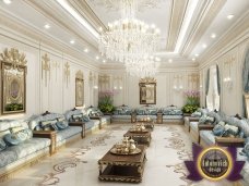 This picture shows a luxurious living room designed with ornate accents. The walls are adorned with luxurious gold and beige wallpaper, while the floor is composed of beige marble tiles. An intricately carved white sofa is the centerpiece of the room, accompanied by two matching armchairs and a leather ottoman. There is also an oval mirror above the sofa, two small end tables near it, and a decorative chandelier hanging from the ceiling. The overall effect is one of opulence and grandeur.