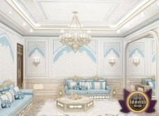 This picture appears to show an ultra-luxury living room with a beige velvet sofa, two gold and white armchairs, a wooden accent table, and crystal chandelier. The walls are painted in a soft beige color, while the chic rug and pillows on the sofa add a touch of elegance and style.