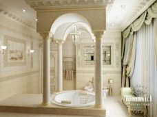 This picture shows a luxurious bathroom with marble flooring, a round spa bathtub surrounded by plants, two sinks, and a standing shower. There is a window with a view of the outdoors, a built-in bench for seating, and a vanity with a mirror and storage. The walls are white and there is an intricate tile pattern along the wall behind the tub and shower.