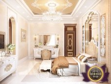 This picture shows a luxurious bedroom with a white and gold decor. The room has a large bed with a white leather tufted headboard, two matching nightstands with white marble tops, two decorative gold chairs, and a large ornate gold framed mirror. The floors are a light parquet and the walls are painted a warm cream color. The ceiling features an elegant crystal chandelier and several recessed lights.