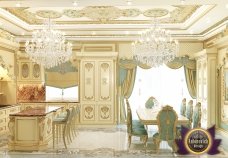 The picture shows a modern, luxurious interior design. It features an elegant marble staircase and railing with ornate gold accents, a white marble floor, and a grand crystal chandelier with multiple tiers. There is a plush tufted sofa in pale pink upholstery and a glass side table with a plant placed on top of it. A large decorative mirror is also present on the wall, adding more light to the room.