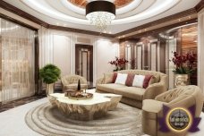 This picture shows a luxurious living room area that includes a unique white color scheme with gold accents. The area consists of two large sofas, a coffee table, and shelves filled with books and decorations. The walls have a custom wallpaper motif, while the floor is covered with a beige-colored rug. The overall effect is one of sophistication and luxury.