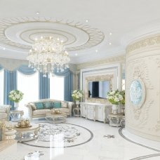 This picture shows a luxury living room with white walls, a white sectional sofa, and a round glass coffee table. The floor is marble and the ceiling has decorative crown molding. There are two chandeliers on either side of the room and a pair of large windows that overlook a scenic view. The decor includes gold contemporary decorations and an art piece hanging on the wall.