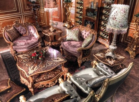This picture shows a grand and elegant living room with rich golden and white tones. The walls are a bright off-white color, adorned with an intricate and elaborate patterned wallpaper in gold and cream. A large beige sofa with matching armchairs and pillows takes up most of one wall, while a large white table with two ornate chairs sits in the center of the room. Above the sofa hang two large sconces with intricate gold detailing, while a large gold framed mirror hangs above the fireplace. There is a white marble floor with a large tan and gold Oriental-style rug