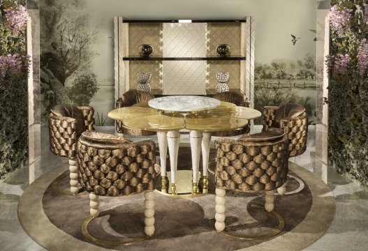 This picture shows a meticulously designed formal dining room. The room is dominated by a large, round white marble dining table with a crystal chandelier suspended above it. The walls are paneled with marble and dark stained hardwood millwork, providing a luxurious and warm feeling to the space. The seating of the room consists of matching chairs upholstered in a light blue and gold fabric. Several decorative elements such as an ornamental mirror and gilded wall accents provide additional elegance to the room.