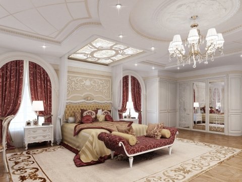 The picture shows a large and luxurious bedroom with a grand bed in the center of the room. Surrounding the bed is an elegant upholstered headboard with golden accents, as well as two matching nightstands and lamps on either side of the bed. The walls are a light blue color, while the window is framed by heavy curtains in a darker blue color. At the foot of the bed is a comfortable sitting area with two armchairs, a coffee table and a round ottoman. Overall, the room has a luxurious and sophisticated feel to it.